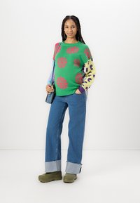 Πράσινο πλεκτό πουλόβερ με ροζ και μωβ λουλουδάτα σχέδια, συνδυασμένο με μπλε ψηλόμεσες τζιν παντελόνες που διαθέτουν αναδιπλούμενη έλευση και πράσινα χοντρά παπούτσια.