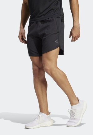 Mężczyzna w czarnych szortach Adidas i białych sportowych butach stoi z jedną nogą lekko wysuniętą do przodu na jednolitym tle.