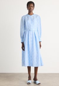 Light blue dress with white polka dots, long sleeves, button front, cinched waist with matching belt, knee-length. Worn with light blue slides.