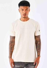 Camiseta de color beige claro con mangas cortas, cuello redondo y logo en relieve en el pecho. Presenta una textura sutil y un diseño ajustado.