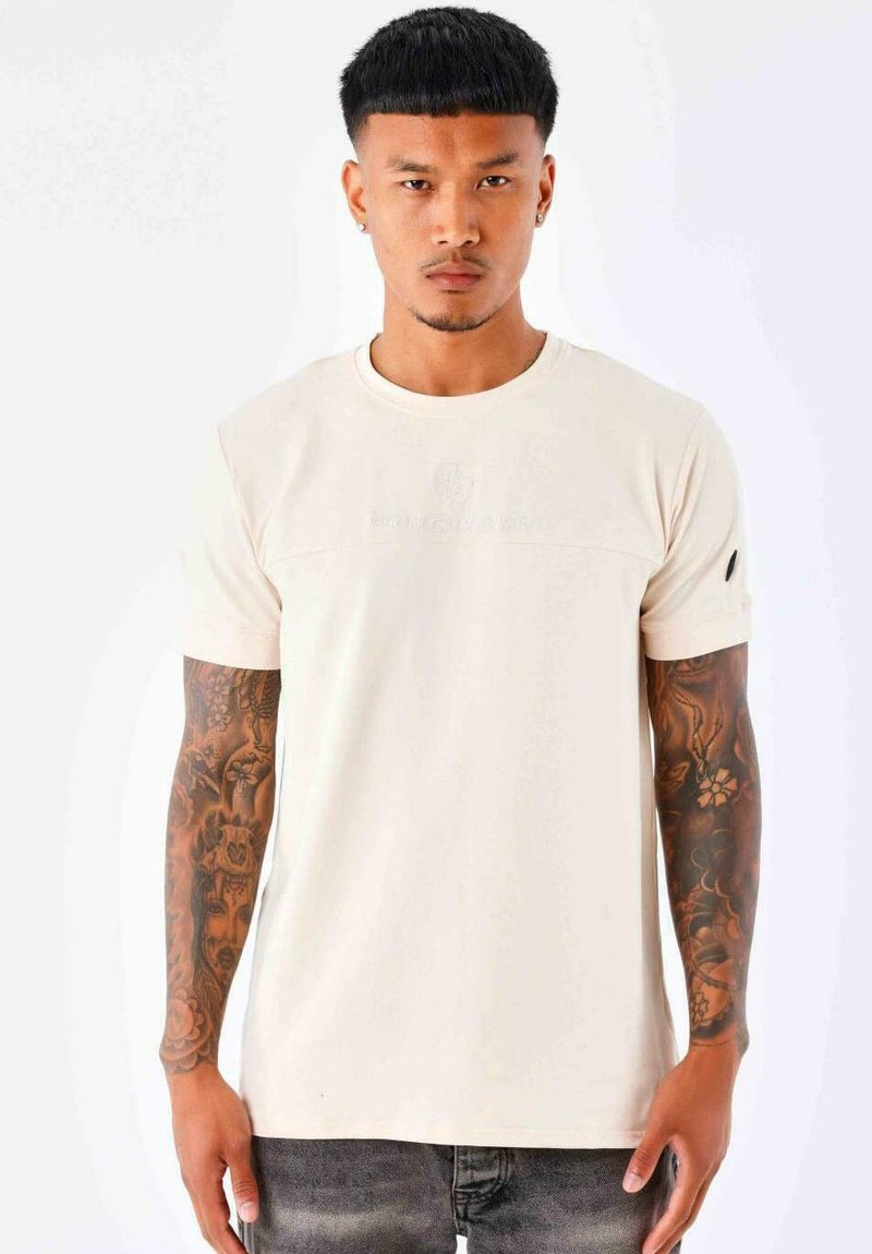 Camiseta de color beige claro con mangas cortas, cuello redondo y logo en relieve en el pecho. Presenta una textura sutil y un diseño ajustado.