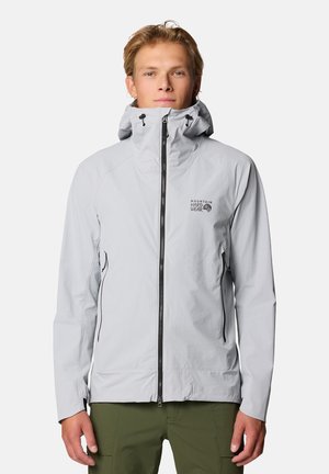 Giovane uomo che indossa una giacca grigio chiaro con zip e cappuccio e pantaloni verde oliva, in piedi davanti a uno sfondo semplice.