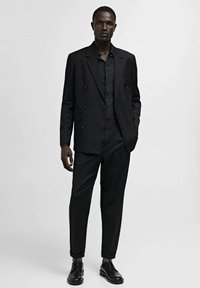 Costum negru cu sacou cu dublă închidere și pantaloni slim-fit, asortat cu cămașă neagră și pantofi de ceremonie negri, pe un fundal neutru.