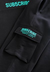 Sort sweatshirt med lys turkise bogstaver: "SUB5CRIBE" på fronten og en label på ærmet med koordinater og "FUTURE SYSTEMS TECHNOLOGY."