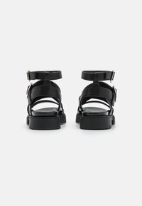 Sandali in pelle nera con cinturini alla caviglia, suola piatta e fibbie regolabili. Presentano un design con punta e tallone aperti, con dettagli di cucitura.
