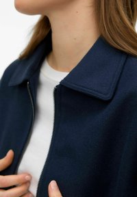 Femme portant un manteau zippé bleu marine par-dessus un haut blanc, visible du cou à la moitié de la poitrine, les mains près de la fermeture éclair.