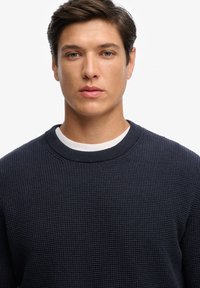 Marineblauwe crewneck trui met textuurgebreide stof, voorzien van een contrasterende witte binnenkraag en geribde manchetten. Close-up van het kledingontwerp.