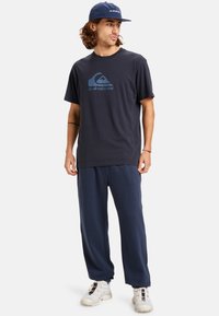 Navy katoenen t-shirt met logo op de borst, gecombineerd met bijpassende navy joggingsbroek. Accessoires zijn een navy pet en witte sportschoenen.