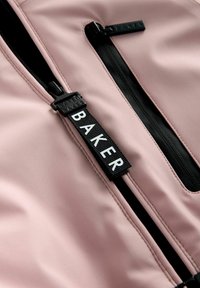 Rozā audums ar gludu tekstūru, kuram ir divi melni rāvējslēdzēji un melna birka ar baltu tekstu "BAKER." R�āvējslēdzējiem ir vilkšanas cilpas.