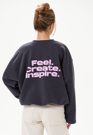 Osoba z włosami związanymi w kok, ubrana w czarną bluzę z różowym napisem „Feel. Create. Inspire.” na plecach i jasne spodnie.