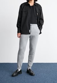 Osoba oblečená v černé košili na knoflíky přes černý top, šedých teplákových kalhotách na šňůrku, bílých ponožkách a černých kožených mokasínech, stojící na modrém koberci.