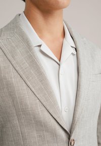 Hellgrauer Leinenblazer mit feinen verticalen Streifen, Revers und Knopfverschluss, kombiniert mit einem weißen Hemd mit Kragen darunter.