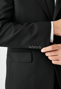 Man die de manchetknoop van een zwart colbert met vier knopen aanpast, over een witte overhemdsmouw.