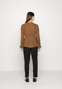 Camisa longa de manga marrom com uma cintura franzida, apresentando um padrão subtil, tecido texturizado e decote redondo. Combinada com calças pretas.