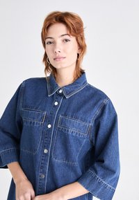 Camisa de mezclilla en azul oscuro, con cierre de botones en la parte delantera, cuello con solapa, dos bolsillos en el pecho y mangas tres cuartos con puños enrollados.