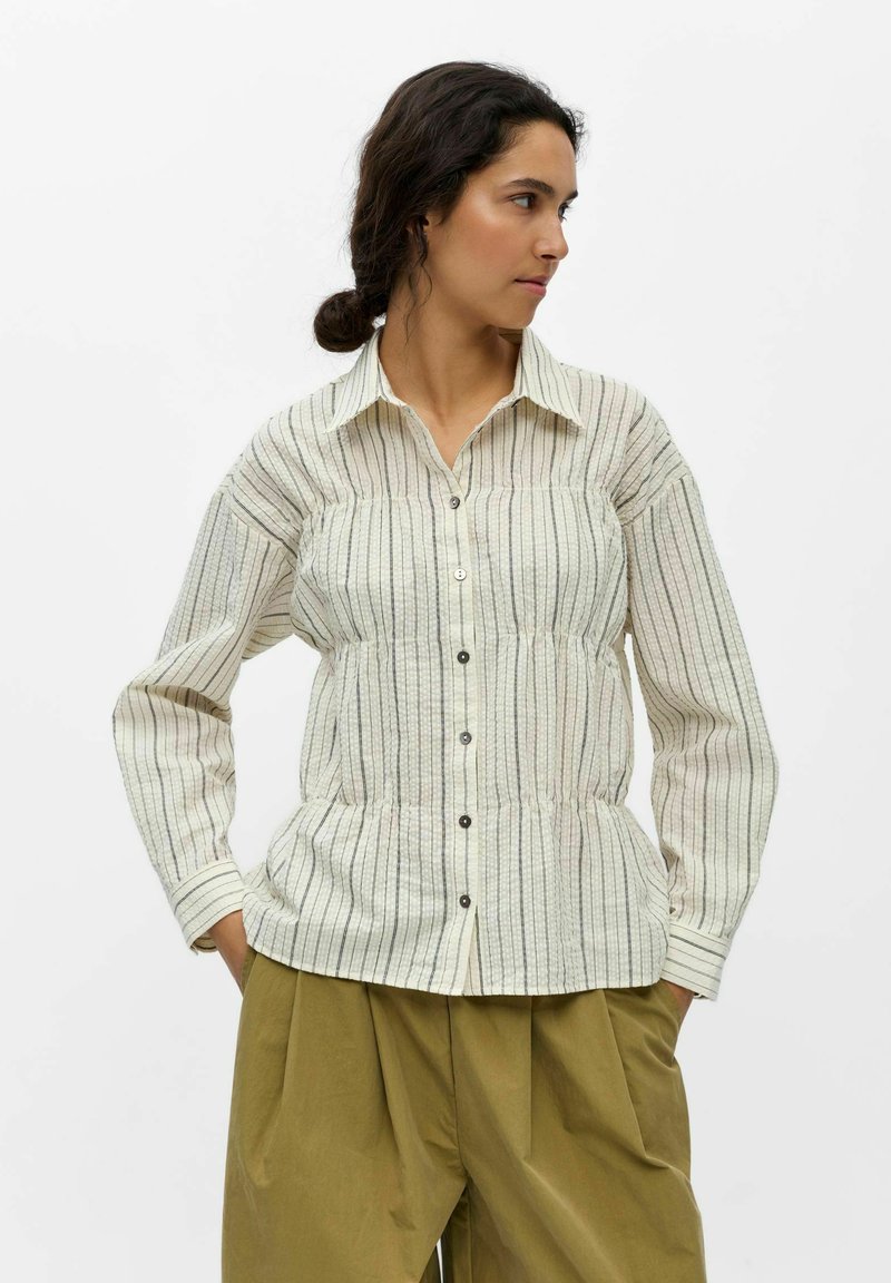 Femme aux cheveux foncés attachés en chignon, portant une chemise beige rayée boutonnée avec une taille cintrée et un pantalon large vert olive sur fond blanc.