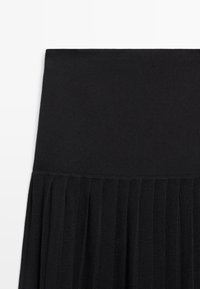 Gonna nera plissettata con una cintura ampia e liscia. Realizzata in un tessuto morbido, presenta pieghe verticali per tutta la parte inferiore.