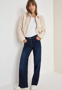 Chaqueta de forro polar crema con cierre de botones, combinada con vaqueros de pierna ancha azul oscuro, que cuentan con un cierre de cinco botones y costuras en contraste. camiseta blanca debajo.