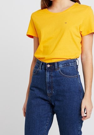 Person trägt ein schlichtes gelbes T-Shirt mit kurzen Ärmeln, das in hochtaillierte blaue Jeans mit Fronttaschen und Gürtelschlaufen gesteckt ist.