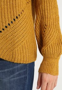 Sennefgelb gestrickter Pullover mit gerippten Bündchen, der diagonale Muster und kleine Löcher an der Seite aufweist. Weiche Textur und chunky Design.