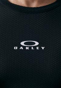 Tricou sport negru cu un model texturat, având un logo alb proeminent "OAKLEY" pe față.