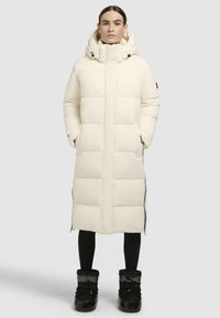 Light cream puffer coat with a hood, featuring large quilted sections, side zippers, and black accents. Worn with black leggings and shiny boots.