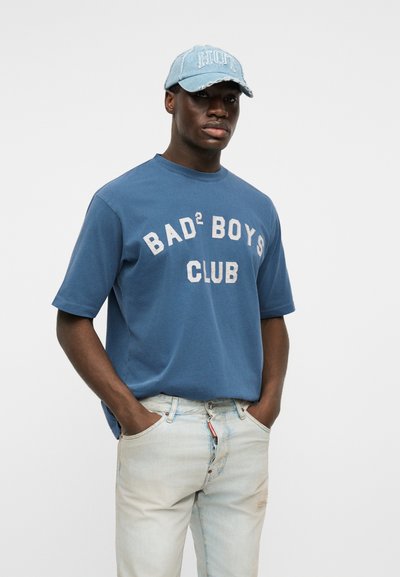 Νέος άντρας με σκουρόχρωμο δέρμα που φοράει μπλε μπλουζάκι με το κείμενο «BAD² BOYS CLUB», ανοιχτόχρωμο τζιν και ανοιχτόγαλανό καπέλο με το κείμενο «HOT», τα χέρια στις τσέπες.