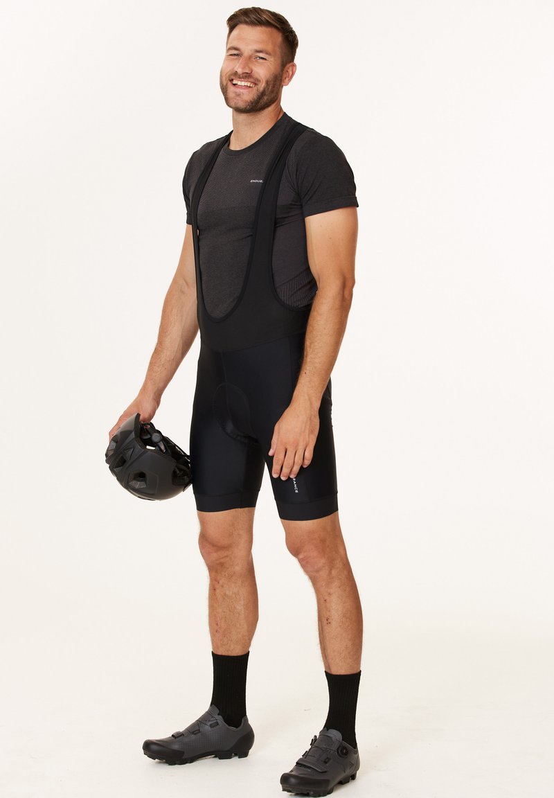 Svarta cykelbyxor med vadderad sits, bärs över en åtsittande grå tröja. Personen håller en svart hjälm och har på sig svarta cykelskor.