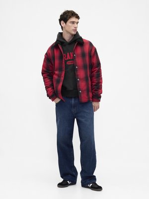 Veste-chemise à carreaux rouge et noir, portée sur un sweat à capuche noir avec logo, associée à un jean bleu ample et des baskets noires.