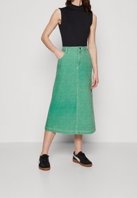 Grön denimkjol med medellång design, två sidofickor och knappstängning; kombinerad med svarta sneakers med vita detaljer.