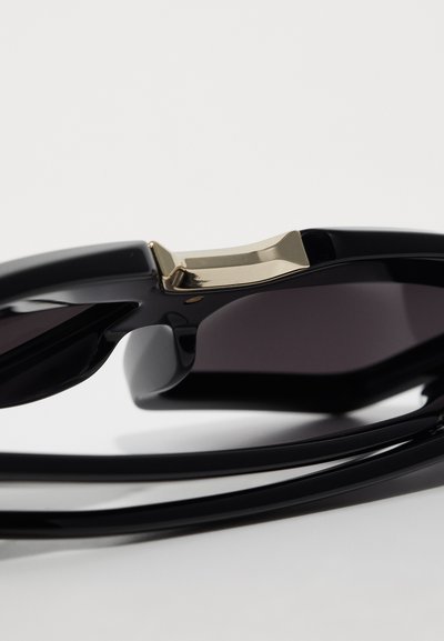 Gafas de sol negras con un acabado brillante, que presentan lentes oscuros y un acento dorado en la patilla del armazón. Diseño elegante y moderno.