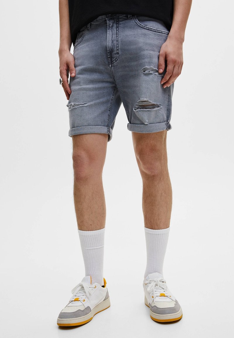 Shorts de denim gris con detalles rasgados, dobladillos enrollados y un ajuste relajado. Combinados con calcetas blancas y zapatillas deportivas con acentos grises y amarillos.
