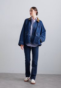 Denim jaka, zila, ar gaišu korduroju apkakli, priekšējām kabatām, brīvs piegriezums, uzvilkta virs gaiša krekla un tumšām džinsām, saskaņota ar sporta apaviem.