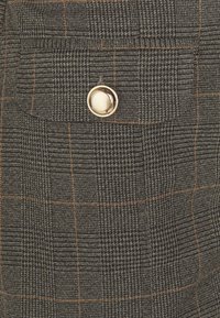 Tessuto a scacchi in tonalità scure con accenti arancioni, caratterizzato da un bottone metallico su una tasca frontale, che mette in evidenza una superficie con texture.