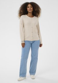 Ženska s kodrastimi lasmi nosi bež kardigan s gumbi, svetlo modre kavbojke, bele superge in srebrno zapestnico, stoji pred enobarvno ozadjem.