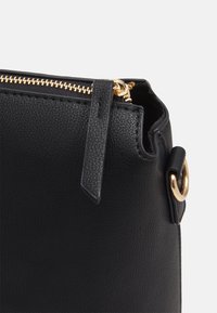 Borsa in pelle nera con superficie testurizzata, zip dorata e un accessorio circolare in oro sul lato. Design compatto, dettagli minimi.