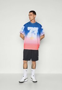Homme debout avec les mains dans le dos, portant un maillot de sport dégradé bleu à rose avec le numéro 73, un short noir, des chaussettes blanches et des baskets blanches.