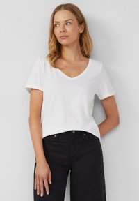 T-shirt blanc à col en V avec des manches courtes roulées, fabriqué en coton doux. Porté avec un jean noir, affichant une coupe décontractée et une texture lisse.