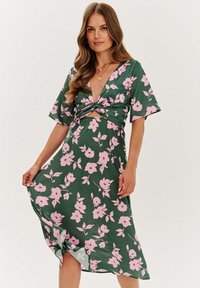 Zelené kvetinové šaty s hlbokým výstrihom do V, krátkymi rukávmi a detailom uzlového pásu. Ozdobené ružovými kvetmi a vzorom listov.