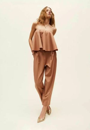 Donna che indossa un top marrone senza spalline e ampio e pantaloni coordinati a vita alta, in piedi con le mani nelle tasche e tacchi metallici dorati a punta.