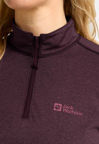 Primo piano di una persona che indossa un pullover Jack Wolfskin viola con zip a quarti, con logo visibile sul petto e capelli biondi.