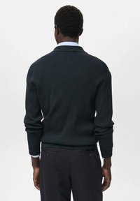 Persona che indossa un maglione a coste scuro e pantaloni scuri, vista da dietro su uno sfondo chiaro e semplice.