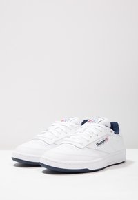 Vita atletiska skor med en slät läderöverdel, perforerad tåbox, blå detaljer på tungan och sulan, med Reebok-logotyp.