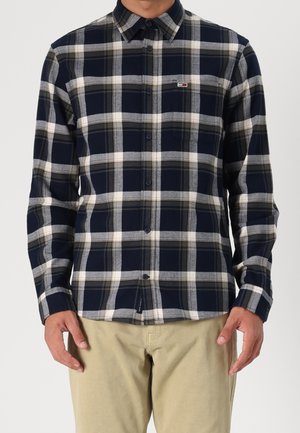 Homme portant une chemise à carreaux bleu marine, blanche et grise, boutonnée avec manches longues, et un pantalon beige, debout devant un fond uni.