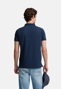 Camisa polo azul marino con cuello, mangas cortas y textura suave, combinada con unos vaqueros de denim claros y una gorra en la mano.