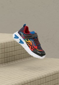 Kinderschuhe mit roten, blauen und orangefarbenen Flammenmustern, schwarzer Kante, Klettverschluss und weißer Sohle, die über geflieste Stufen schwebt.