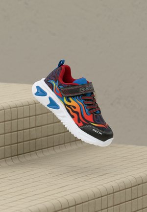 Kindersneakers met rode, blauwe en oranje vlampatronen, zwarte afwerking, klittenband en een witte zool, weergegeven terwijl ze boven betegelde treden zweven.
