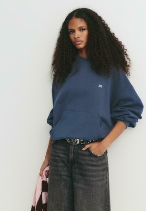 Giovane donna con capelli ricci, indossa un felpa blu navy e jeans neri, tiene accanto a sé un indumento lavorato a maglia rosa e marrone.