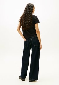 Czarna krótka koszulka, ciemnoniebieskie spodnie z szerokimi nogawkami z kontrastowym przeszyciem i fakturowane czarne obuwie. Włosy są długie i faliste.