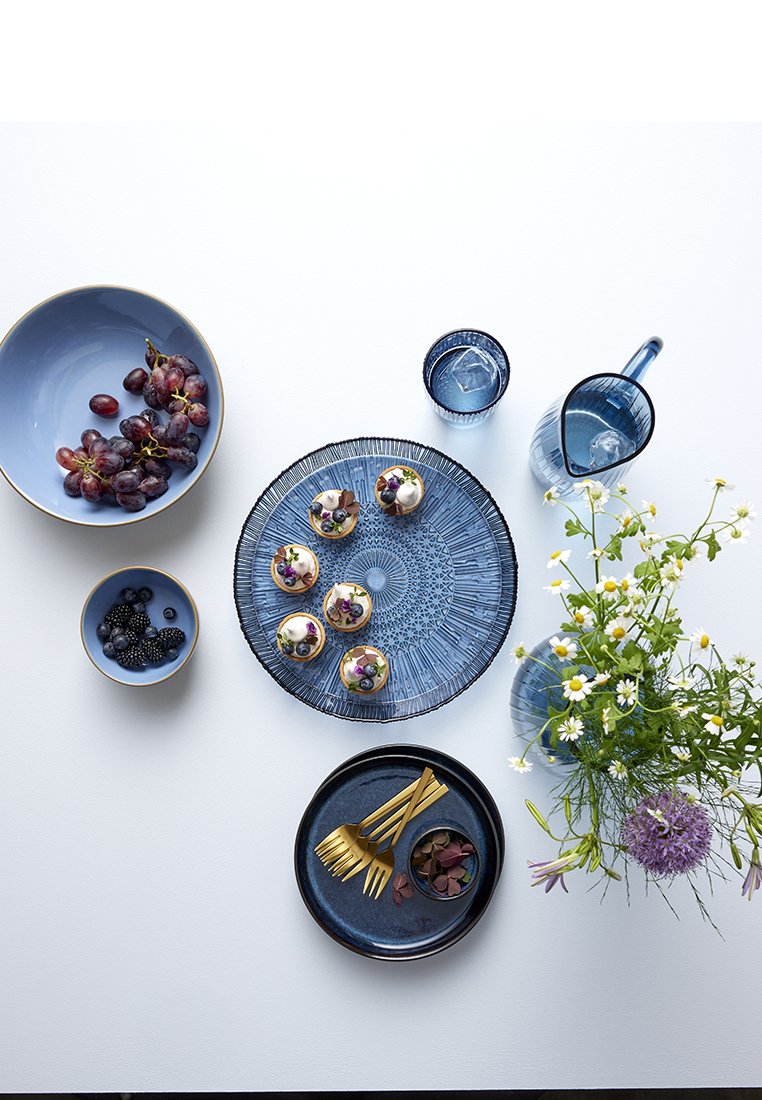 Blaue Keramikteller und -schalen mit strukturierten Mustern, goldenes Besteck, frische Früchte und Blumen, die auf einem weißen Tisch arrangiert sind.